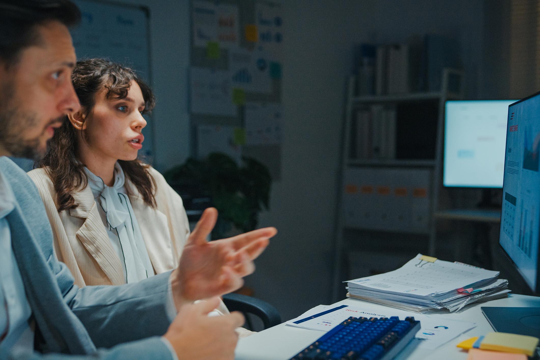 Two coworkers have a late-night business meeting in office, discussing work updates and reviewing information on the computer screen. Teamwork, mentorship, overtime and deadline pressure.